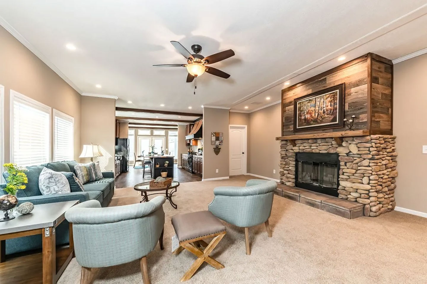 The living room of a manufactured home — a stacked-stone fireplace with a reclaimed-wood surround, open through to the kitchen.